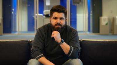 A man with a beard sits on a dark sofa in an office space, resting his chin on his hand. He wears a dark sweater, jeans, and a smartwatch. The background features glass doors and office decor.