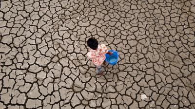 Person holding a blue bucket walking on cracked, dry ground.